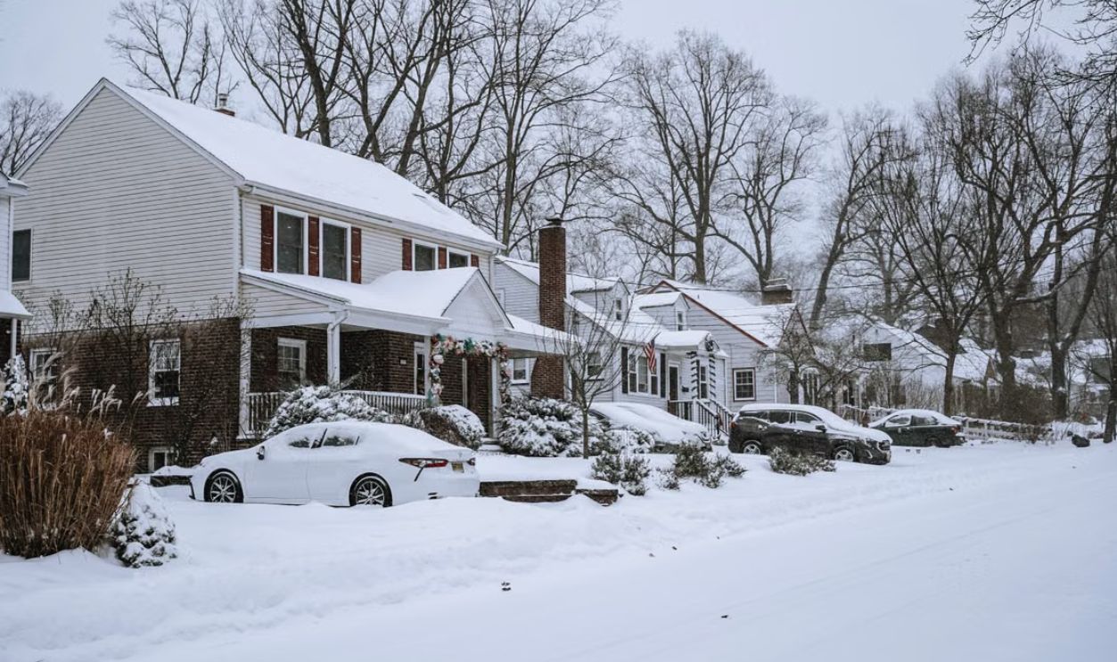 A quiet suburban street blanketed in snow with houses, cars, and leafless trees in winter