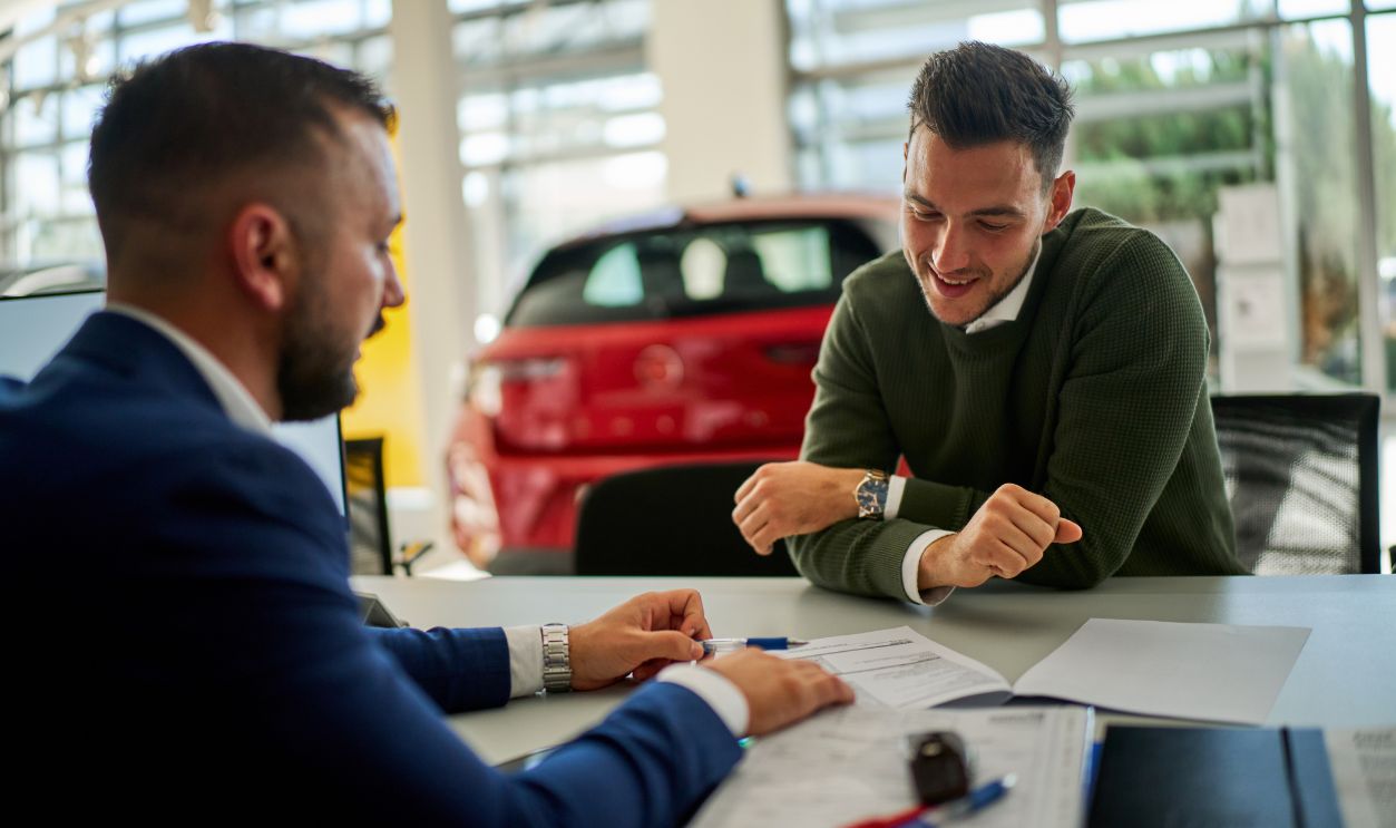 Shutterstock-2582442077, Car salesman and a customer engage in a discussion at a dealership table. Documents and a car key are on the table, with a vehicle visible in the background through large windows