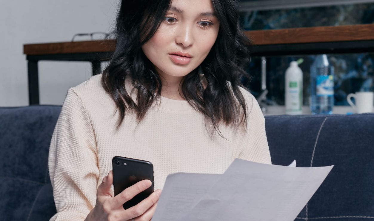 Young woman reviews documents and phone indoors, showcasing concern while seated on a couch with scattered papers.