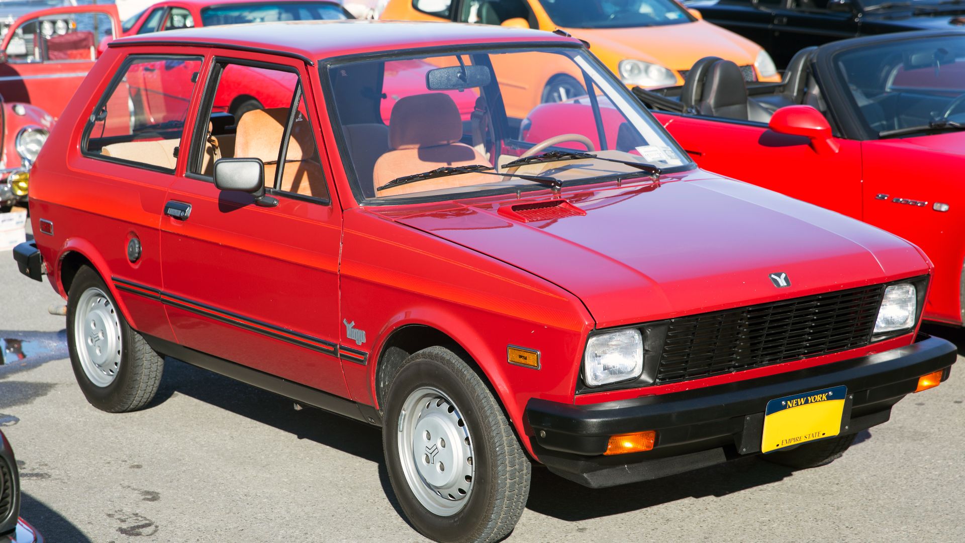1987 Yugo GV in Burgundy with dealer-installed decals and Tan interior, at the wheels_of_nyc Cars & Coffee at the Greenpoint Terminal Market, Brooklyn. 1.1-litre engine and four-speed manual transmission. Originally delivered in Iowa, spent decades in a M
