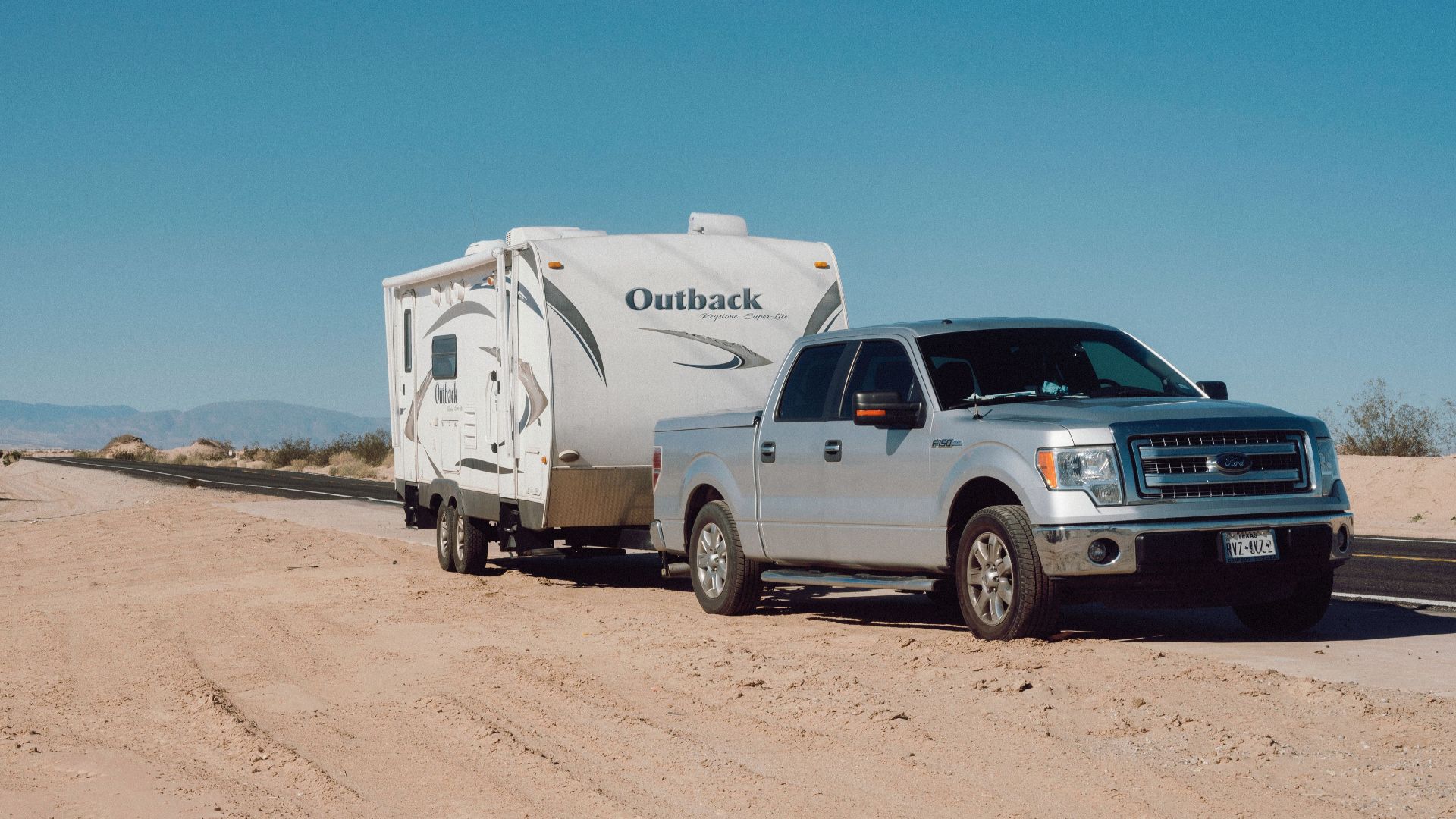 Silver truck towing an Outback trailer on a desert road under clear blue skies.