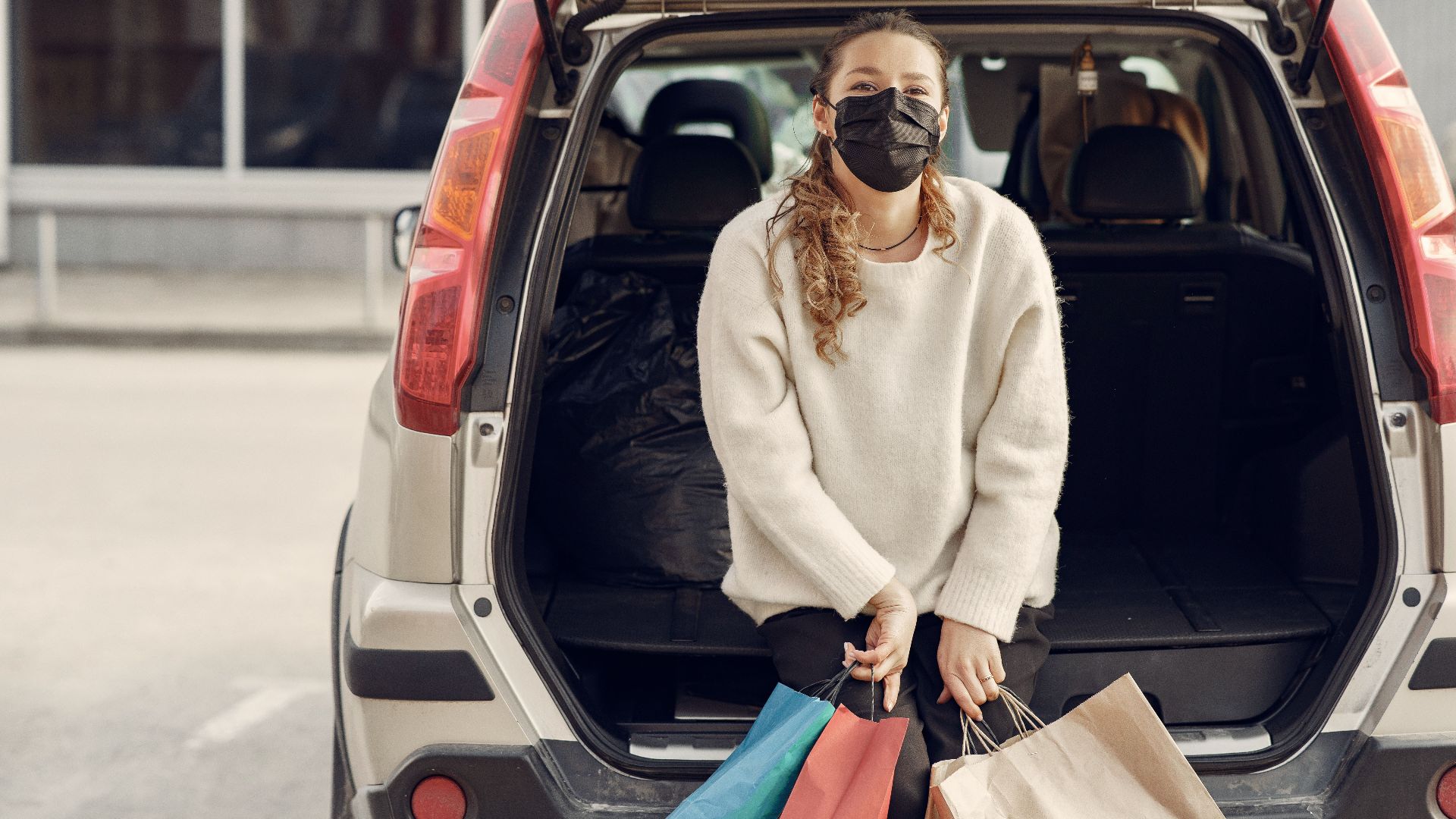 Young woman in casual clothes and protective mask sitting in trunk of modern car holding shopping bags with purchases on city street