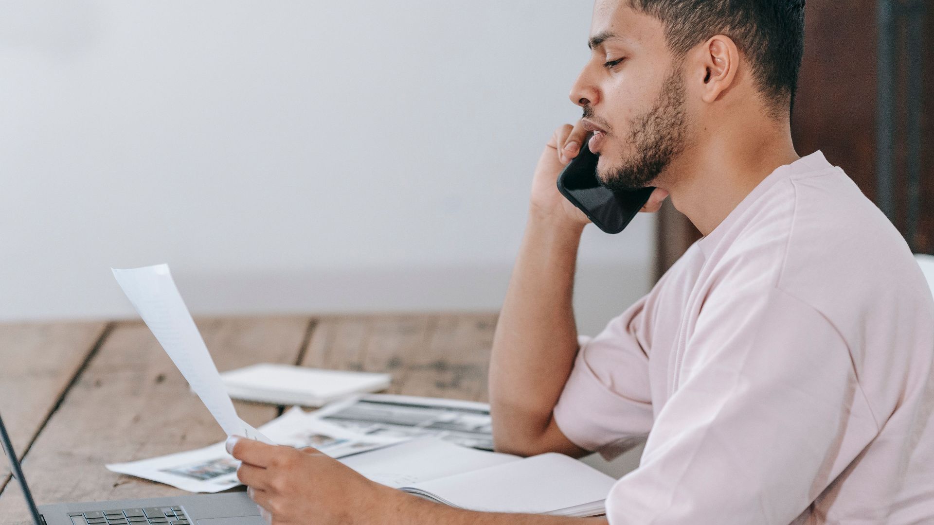 Side view of young Hispanic male remote employee discussing documents during phone call while working online with laptop and papers in home office