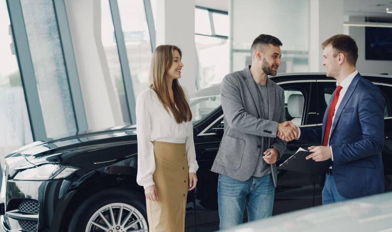 Customers shaking hands with dealer in showroom, sealing car purchase deal.