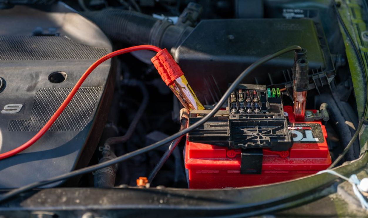 Close-up of a car battery with attached jumper cables in an engine bay.