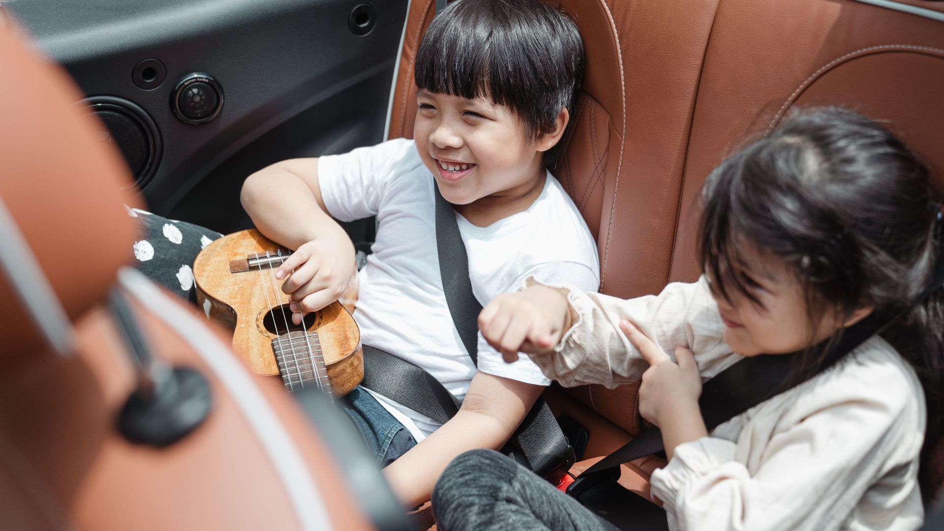 From above smiling ethnic boy and girl in casual outfits sitting fastened in passenger seats with ukulele during road trip together