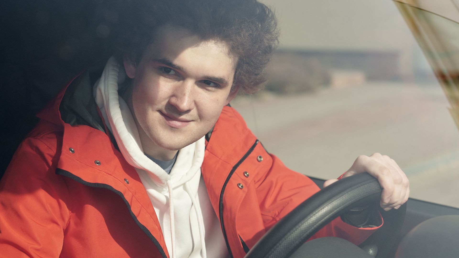 A young adult man smiling while sitting behind the steering wheel of a vehicle.