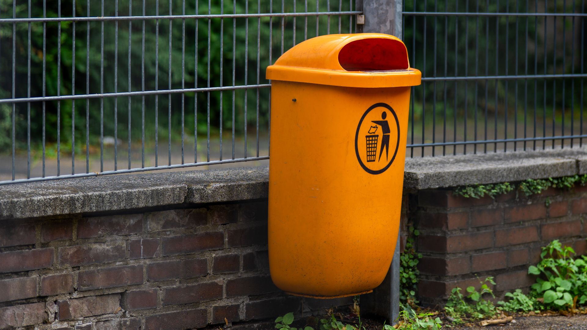 An orange trash bin attached to a brick wall and metal fence, adding contrast to the urban scene.