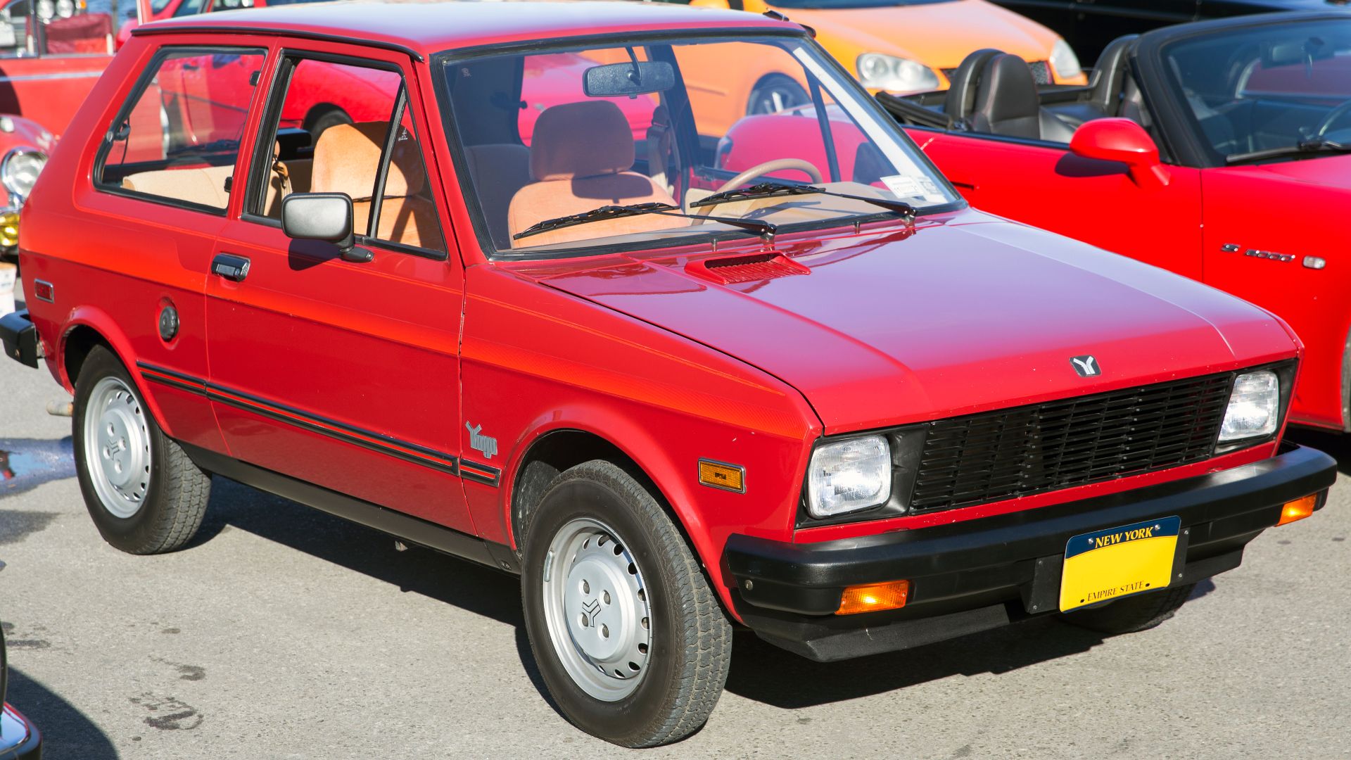 1987 Yugo GV in Burgundy with dealer-installed decals and Tan interior, at the wheels_of_nyc Cars & Coffee at the Greenpoint Terminal Market, Brooklyn. 1.1-litre engine and four-speed manual transmission. Originally delivered in Iowa, spent decades in a M