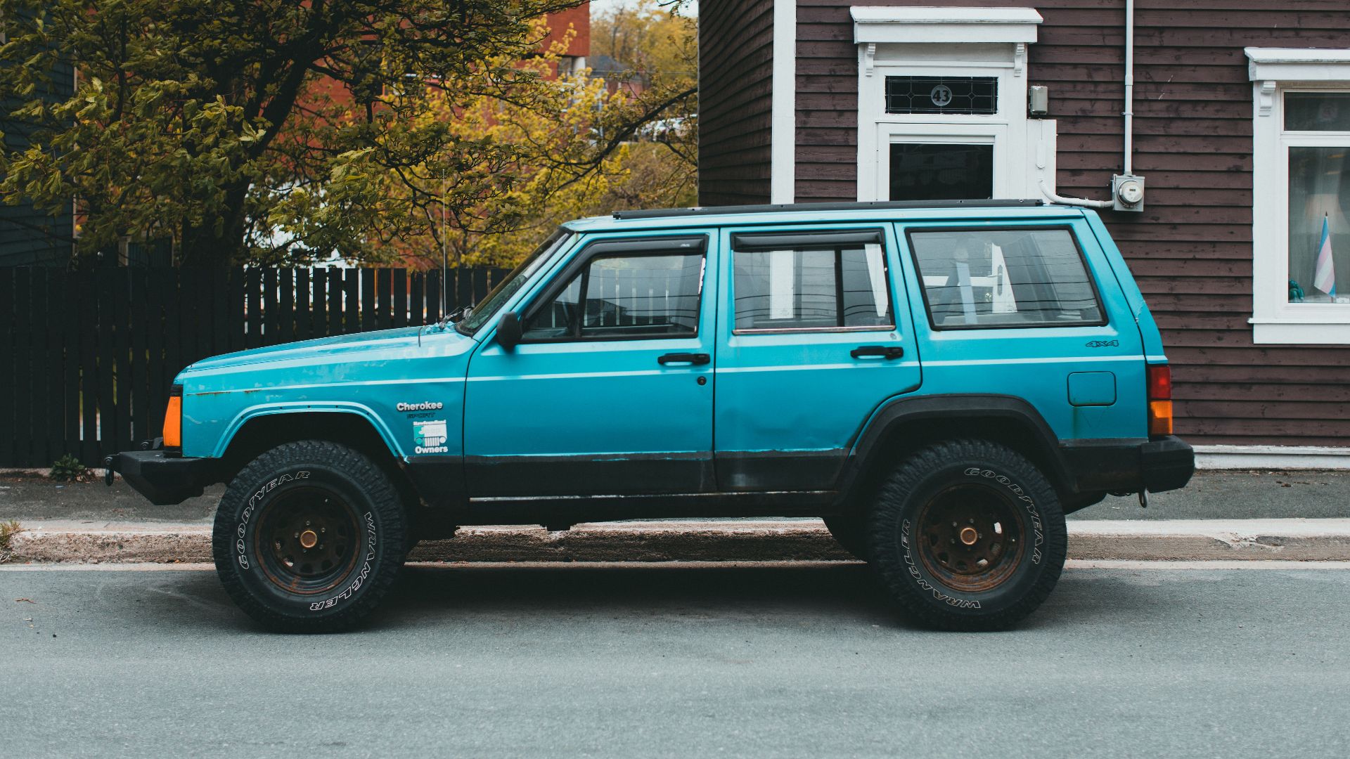 Bright blue vintage Jeep Cherokee parked by a rustic building on a tree-lined street.