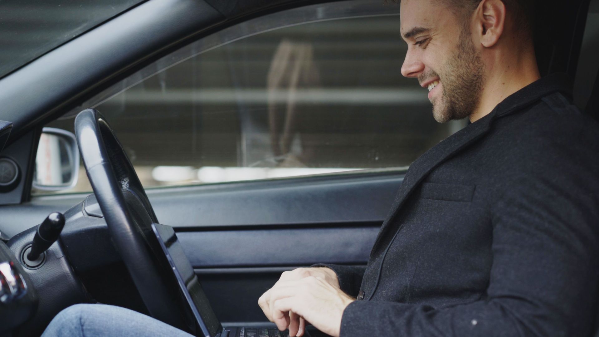A man in casual attire is smiling while working on his laptop inside a car.