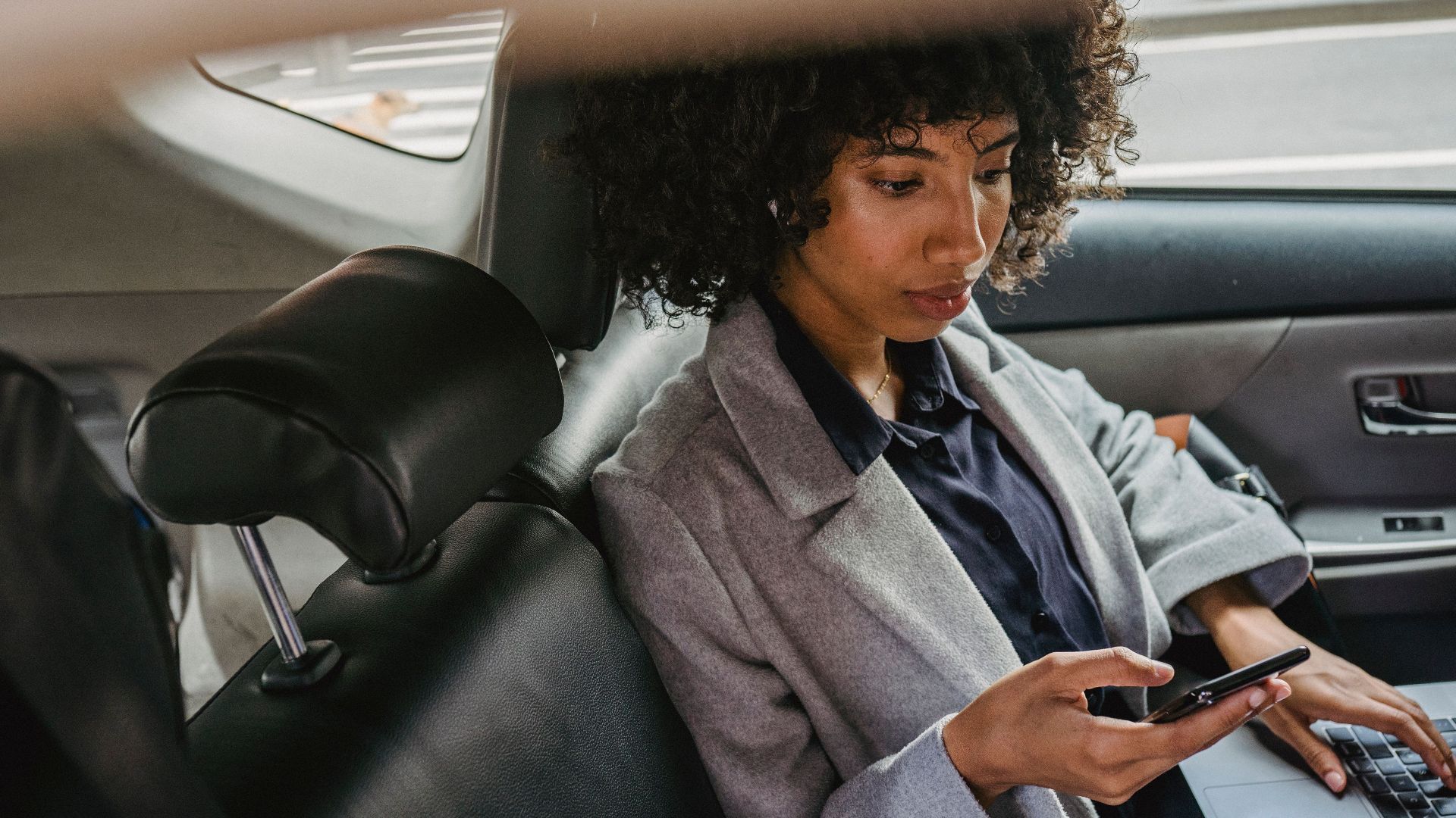 Woman multitasking with phone and laptop in car backseat, balancing work and travel.
