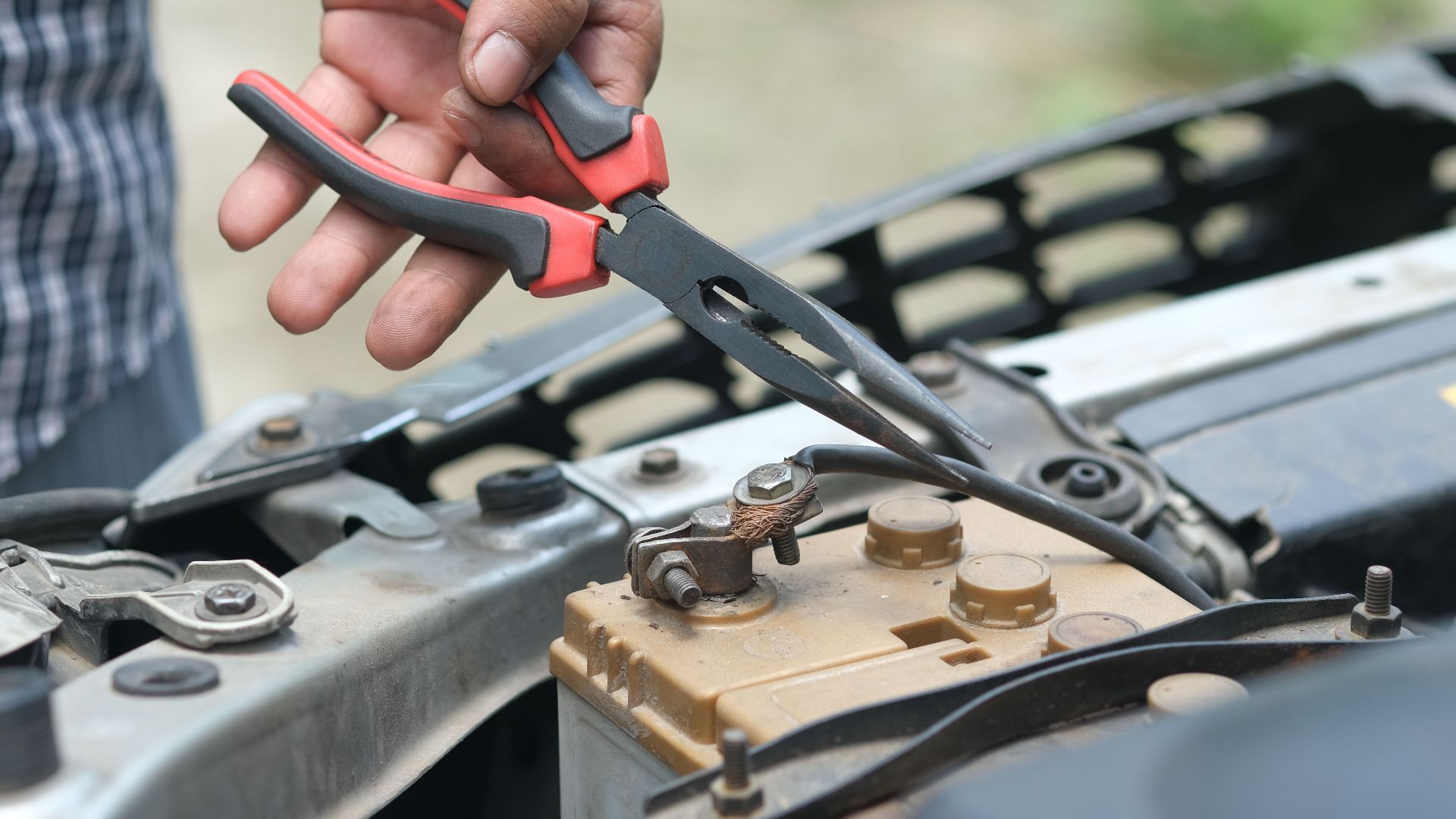 Person using pliers to check a car battery in an outdoor setting.
