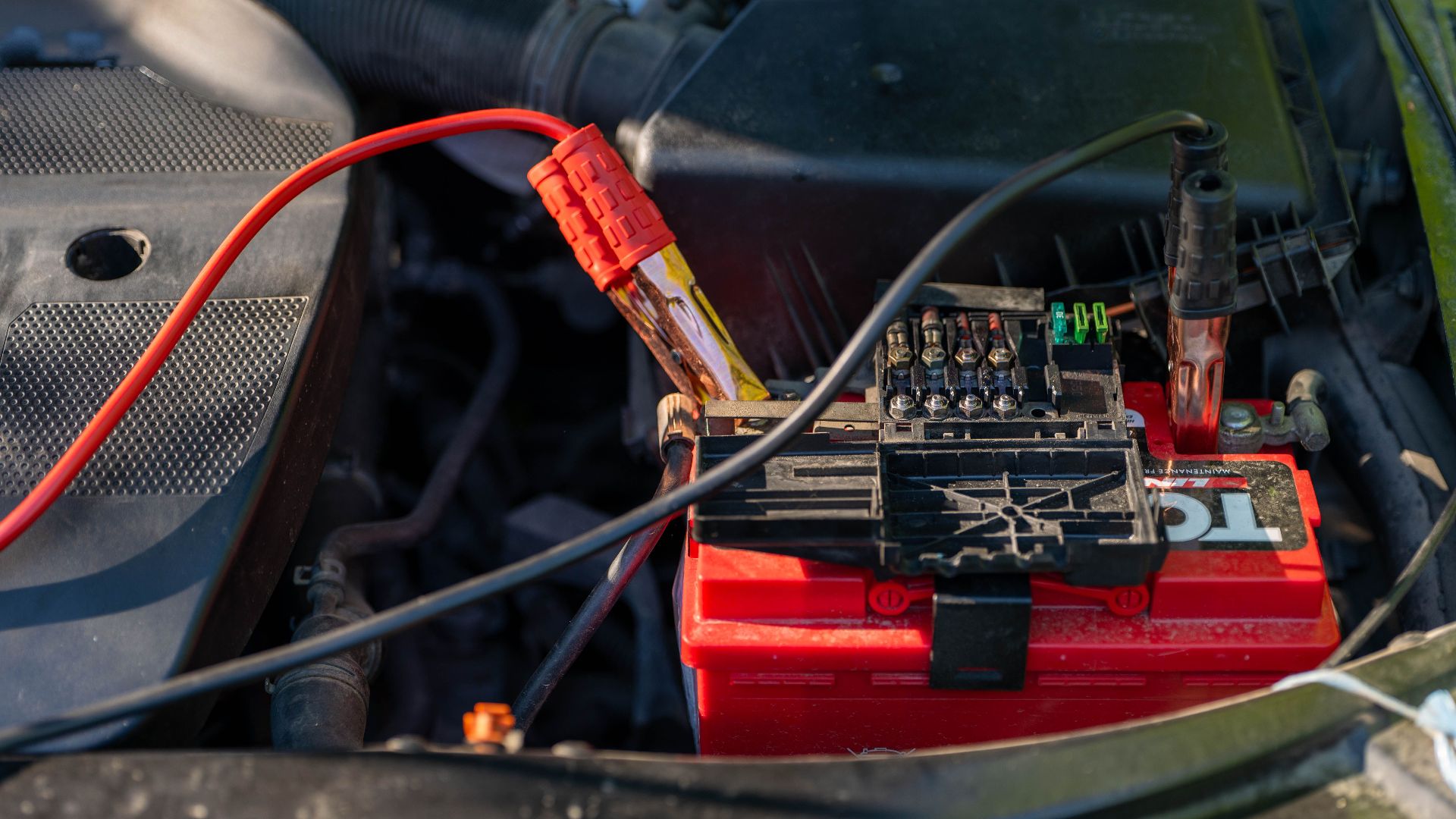 Close-up of a car battery with attached jumper cables in an engine bay.