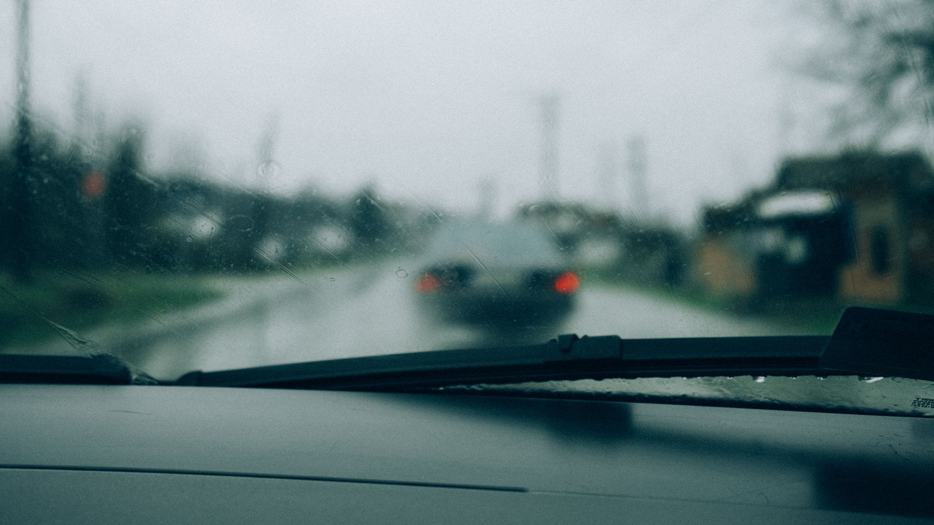 Rain falls steadily on a suburban street, creating a blurred view of a gray car ahead. The scenery reflects a calm yet overcast atmosphere, typical of a rainy day.