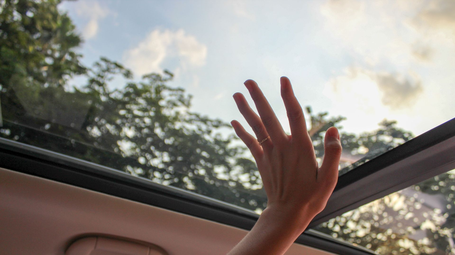 A hand reaching out through a car sunroof, touching the sky on a sunny day.