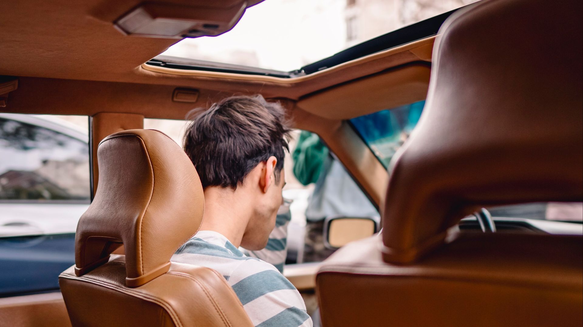 Back view of man sitting inside a car with an open sunroof, viewed from the back seat.
