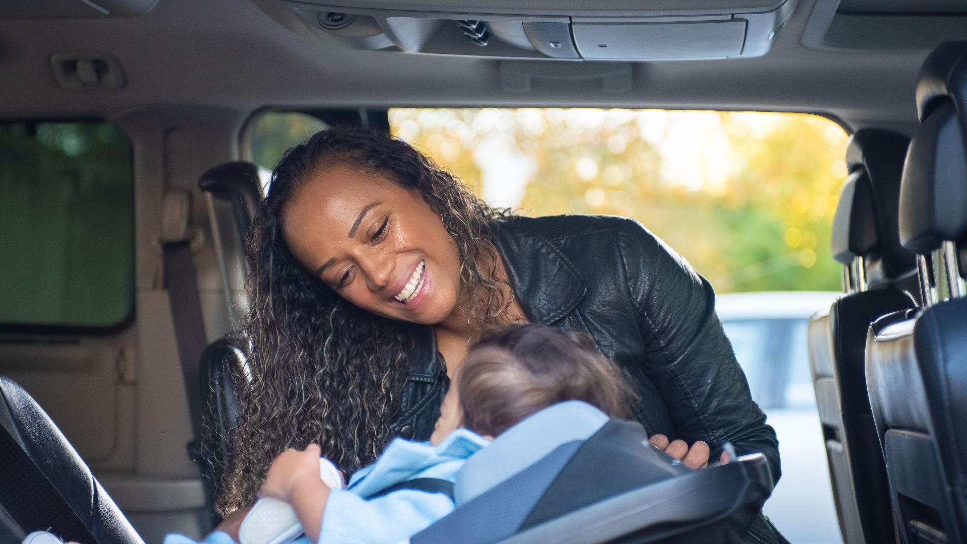 A joyful mother smiles at her baby in a car seat inside a vehicle.
