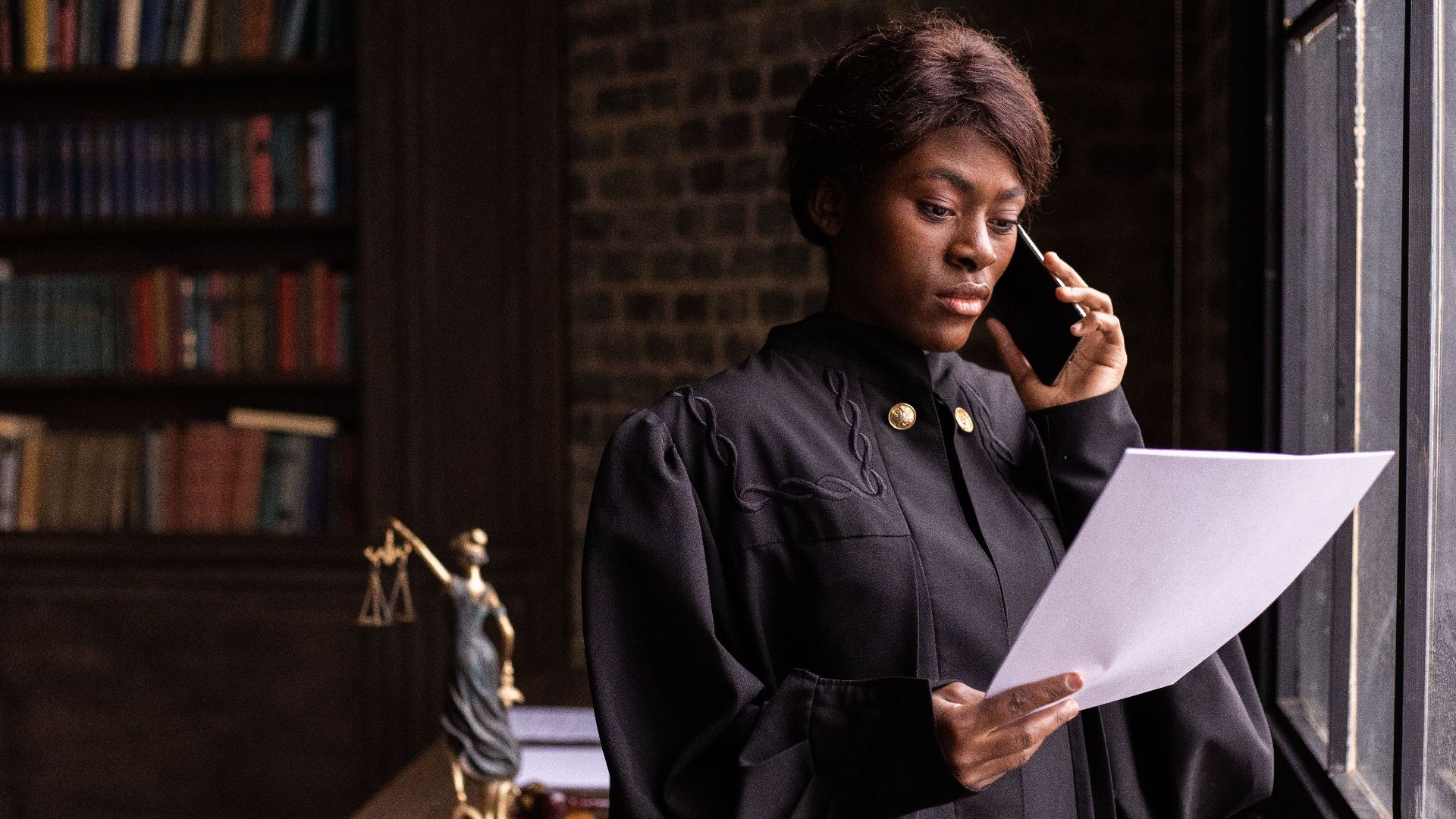 African American judge in robe, reading document and talking on phone by the office window.