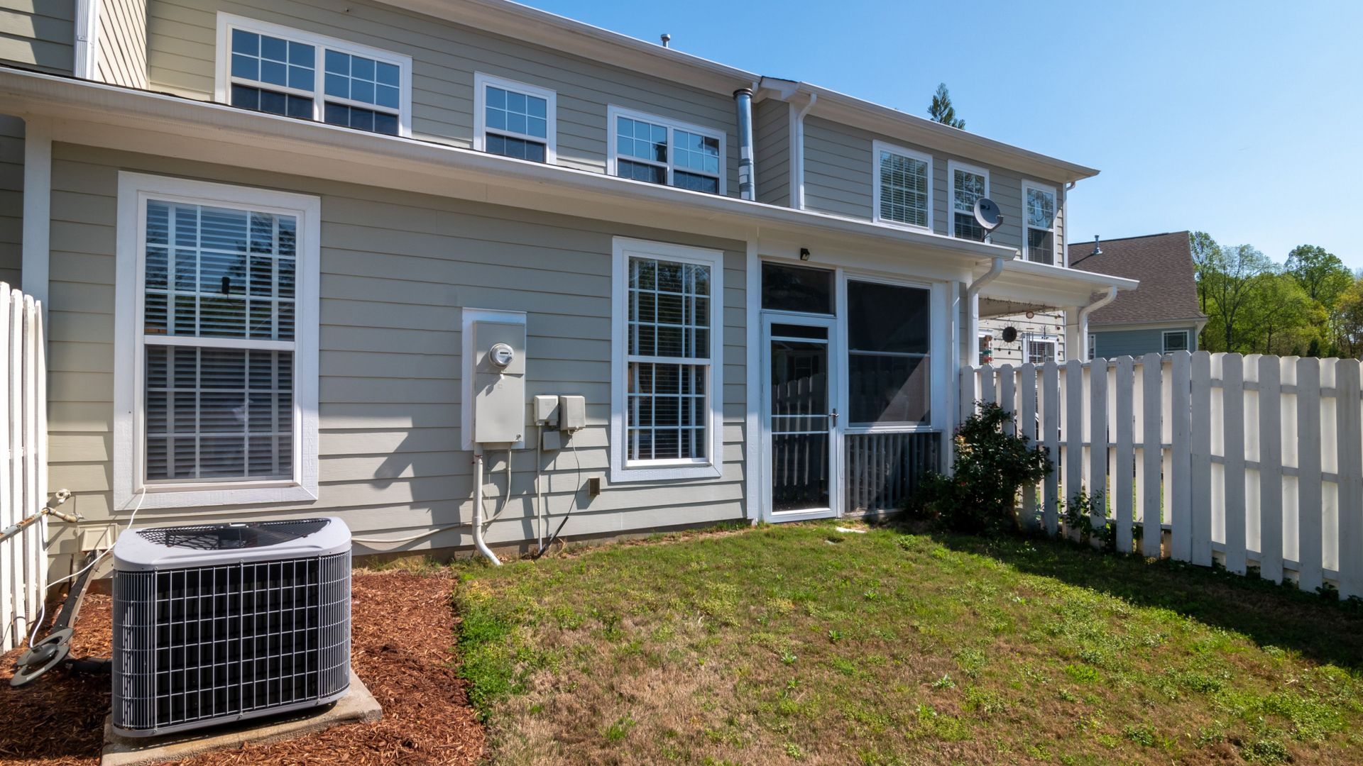 A sunlit backyard view of a modern grey house with white fencing and AC unit.