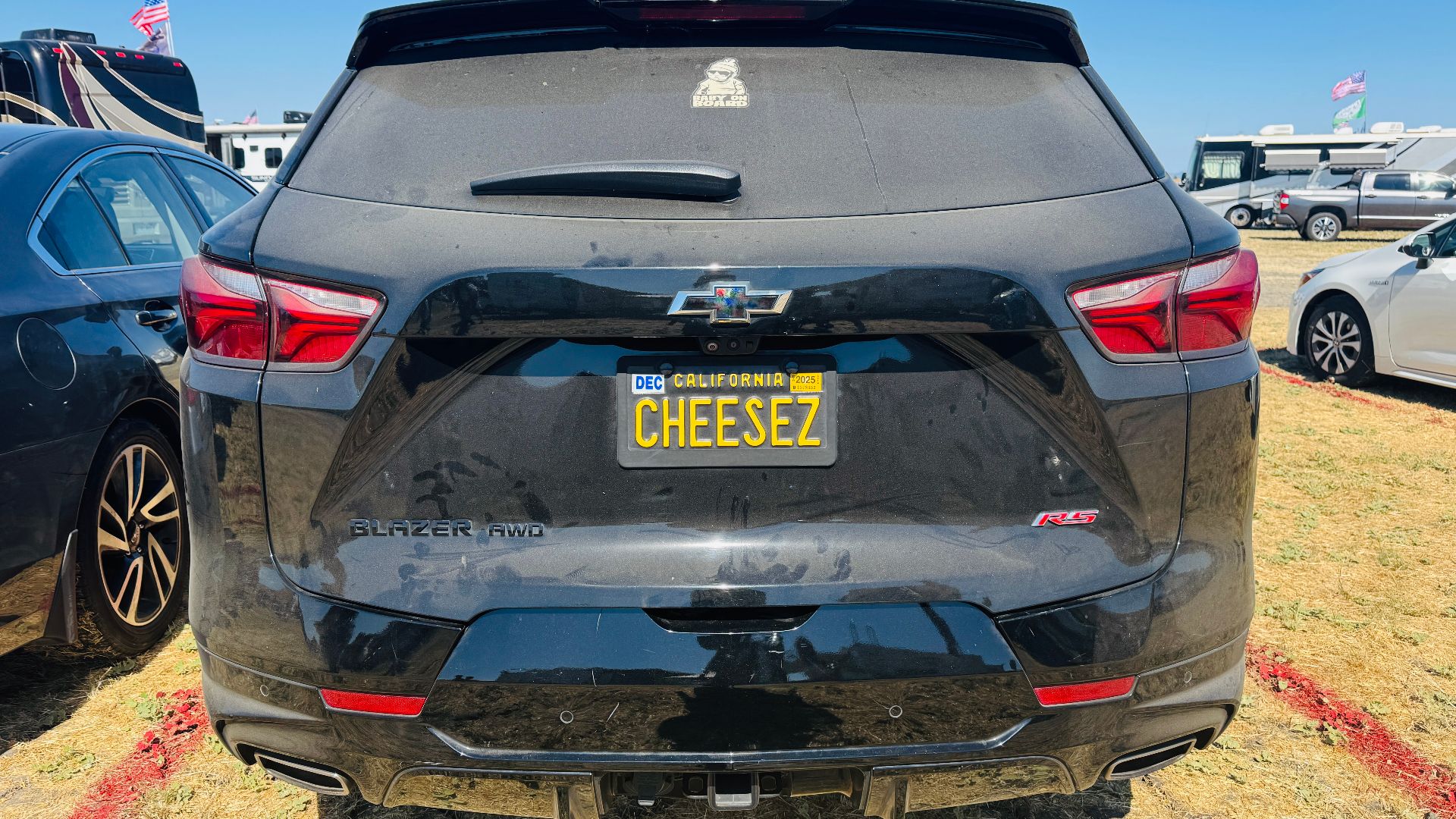 Rear view of a dusty black Chevrolet Blazer RS AWD with a California vanity license plate reading ‘CHEESEZ.’ The SUV is parked on dry grass at an outdoor event, with other vehicles and RVs nearby and American flags visible in the background.