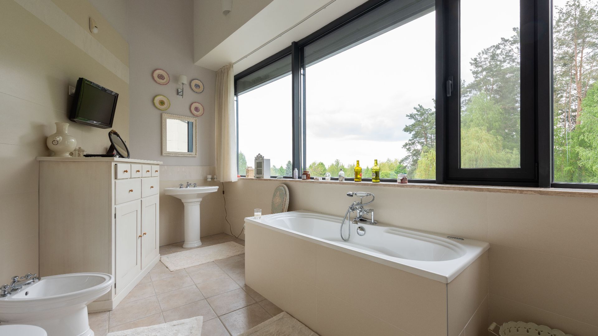 Toilet and bidet near cupboard in stylish bathroom with sink and white tub near big window overlooking green plants in tropical resort