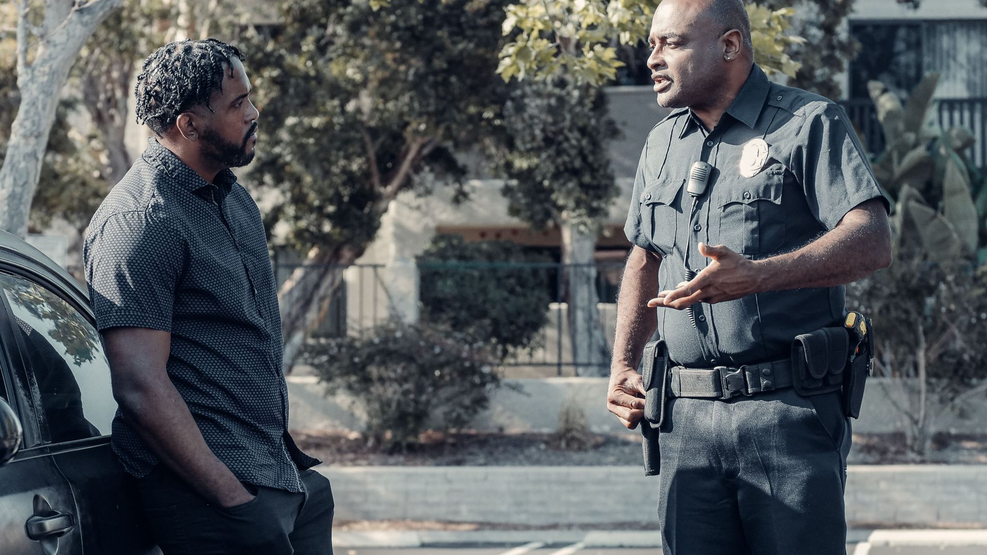 A uniformed police officer converses with a man leaning on a car outdoors.
