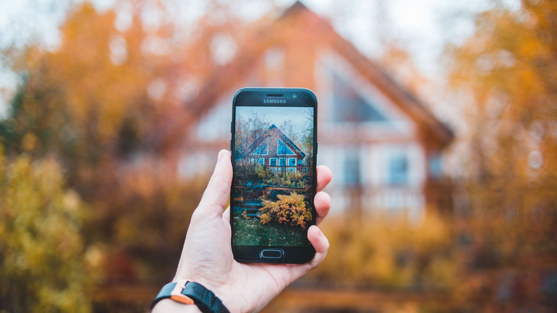 A hand holds a smartphone to photograph a house framed by autumn colors.