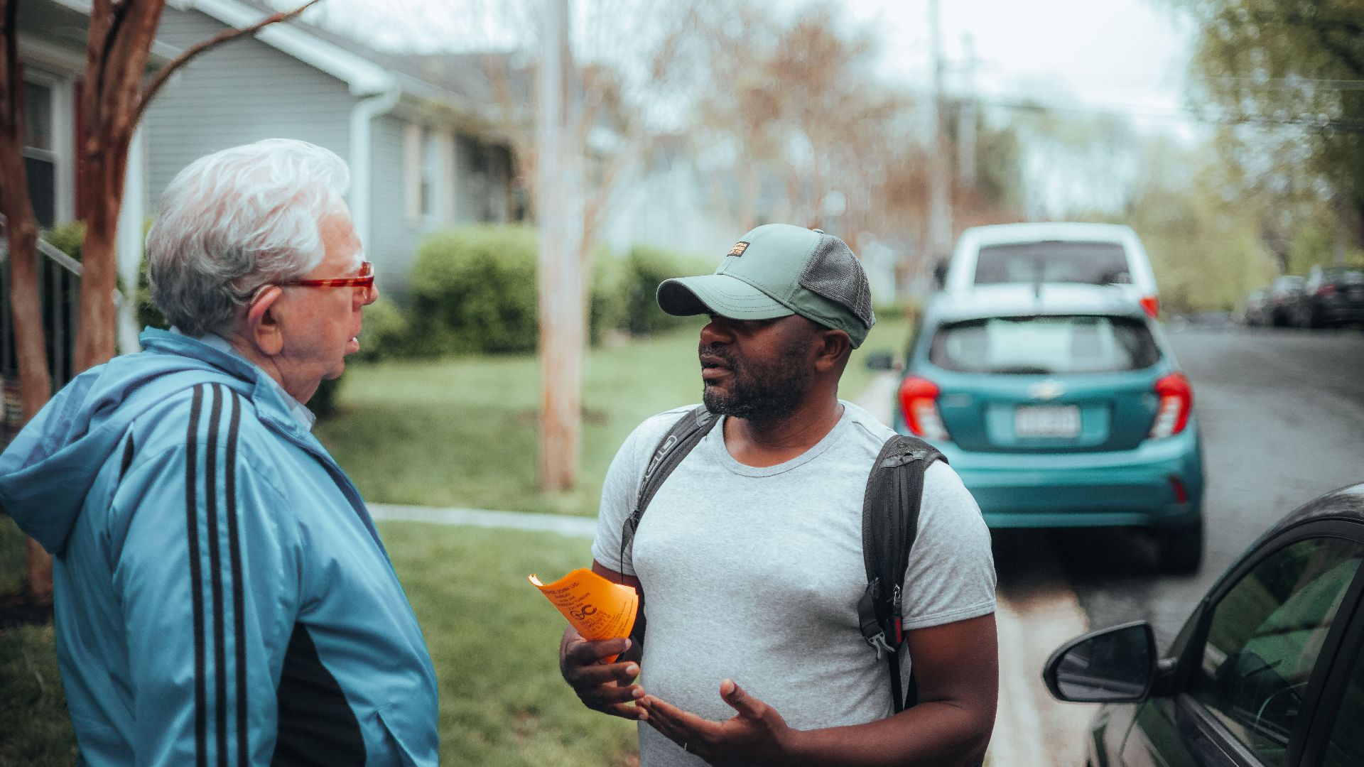 Two men engaged in conversation on a suburban sidewalk with parked cars.
