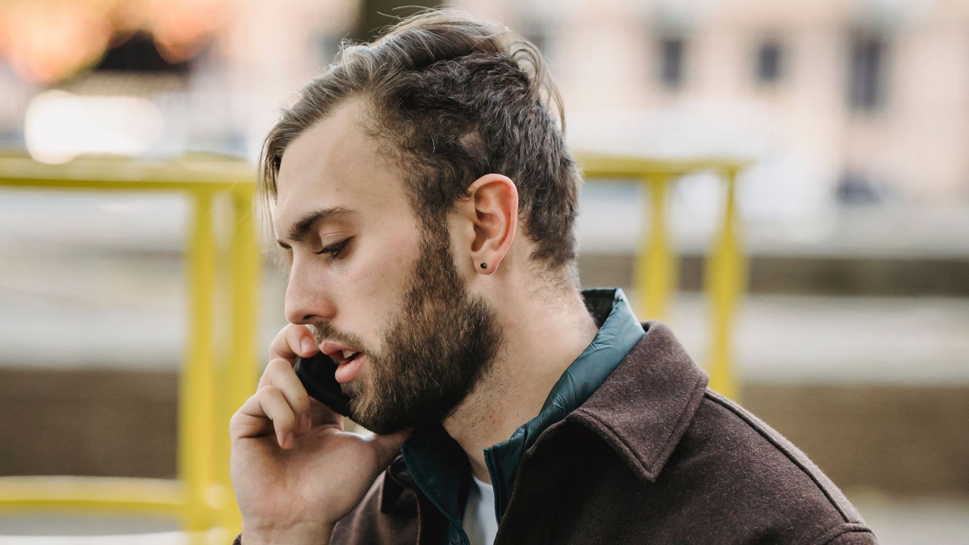 Side view of young bearded man in suede jacket talking on smartphone on playground