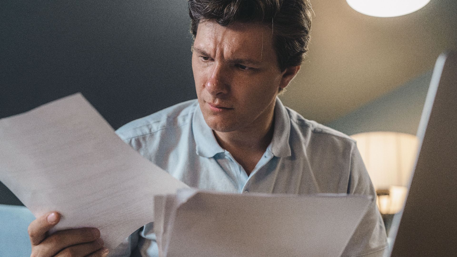 A man intently examines papers, seated indoors under warm lighting, focusing on his work.