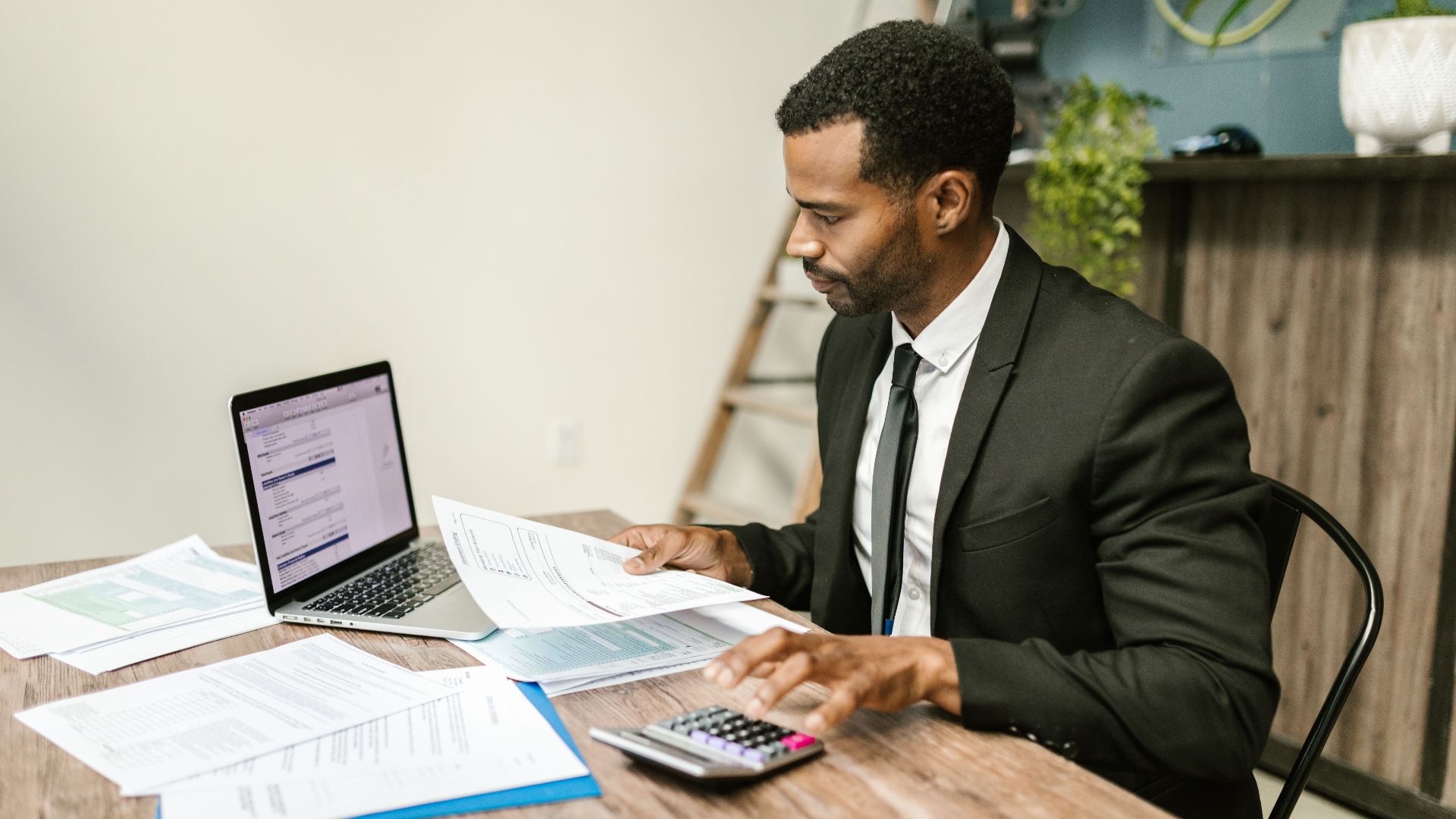 A businessman in a suit working on documents and using a laptop at an office desk.