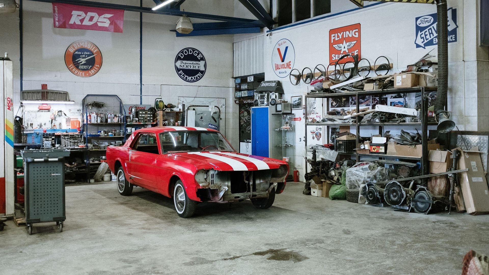A vintage red car undergoing restoration in a well-equipped workshop with various tools and auto parts.