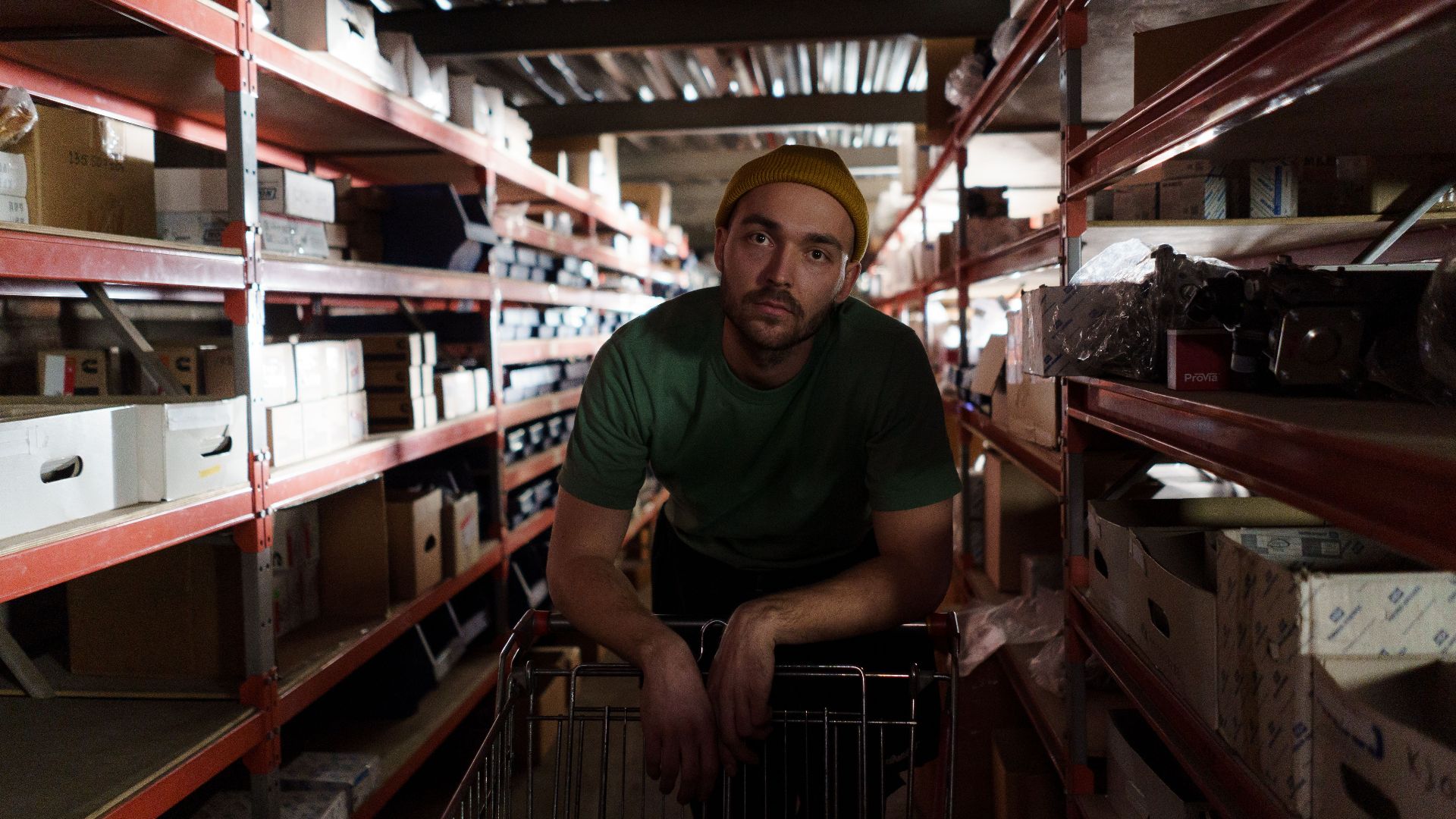 Man in green shirt and beanie pushing a cart in a busy warehouse aisle.