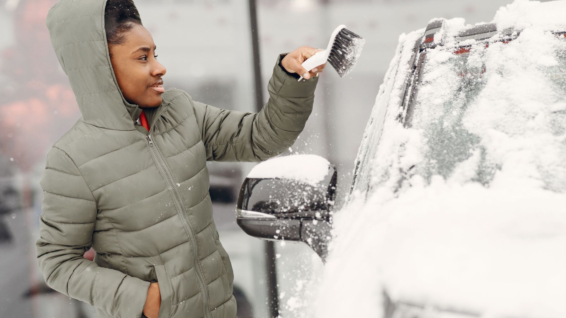 A woman in a green jacket removes snow from her car during winter outdoors.