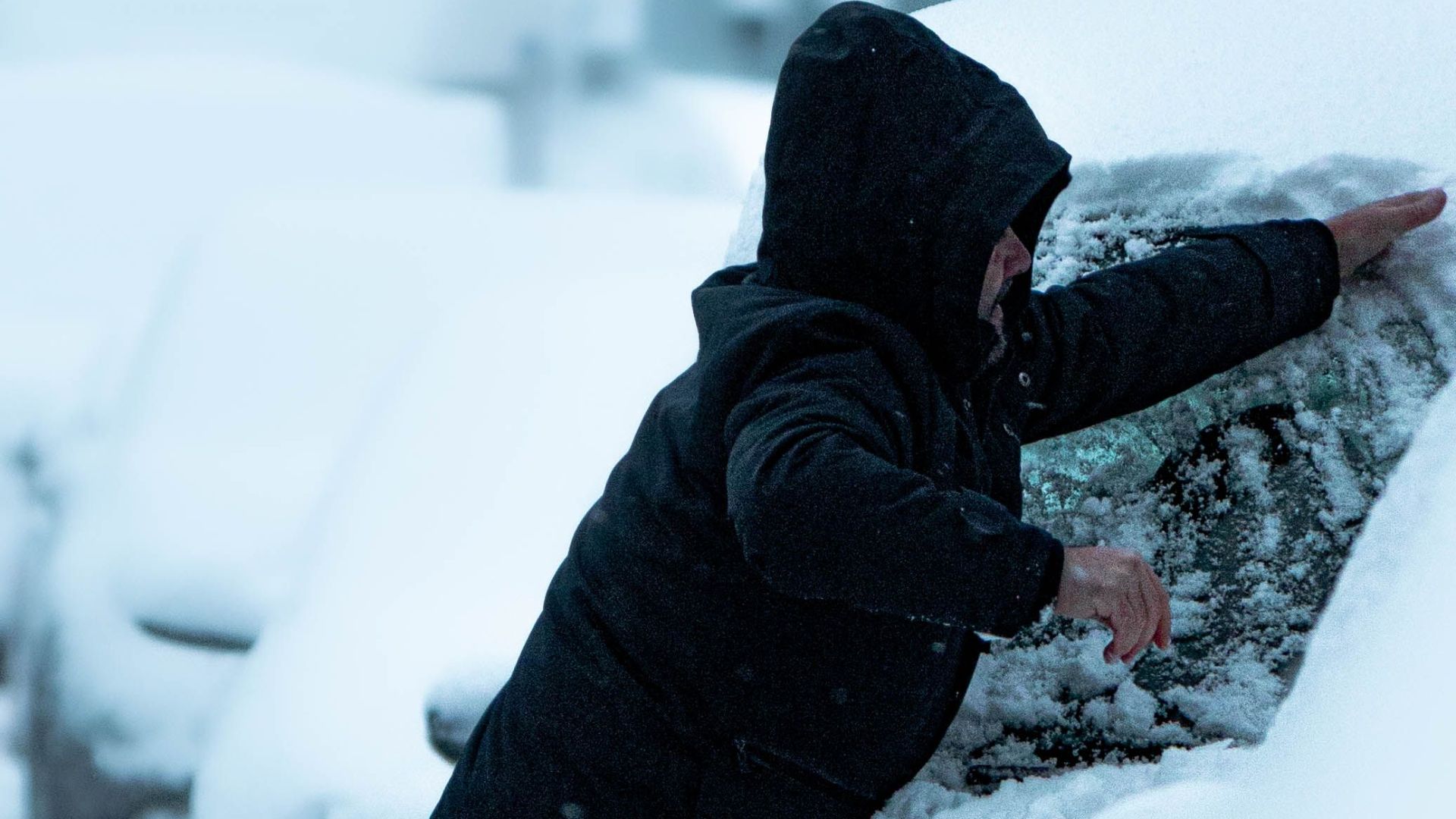 A man clears snow from a car parked on a wintery street, highlighting seasonal car maintenance.