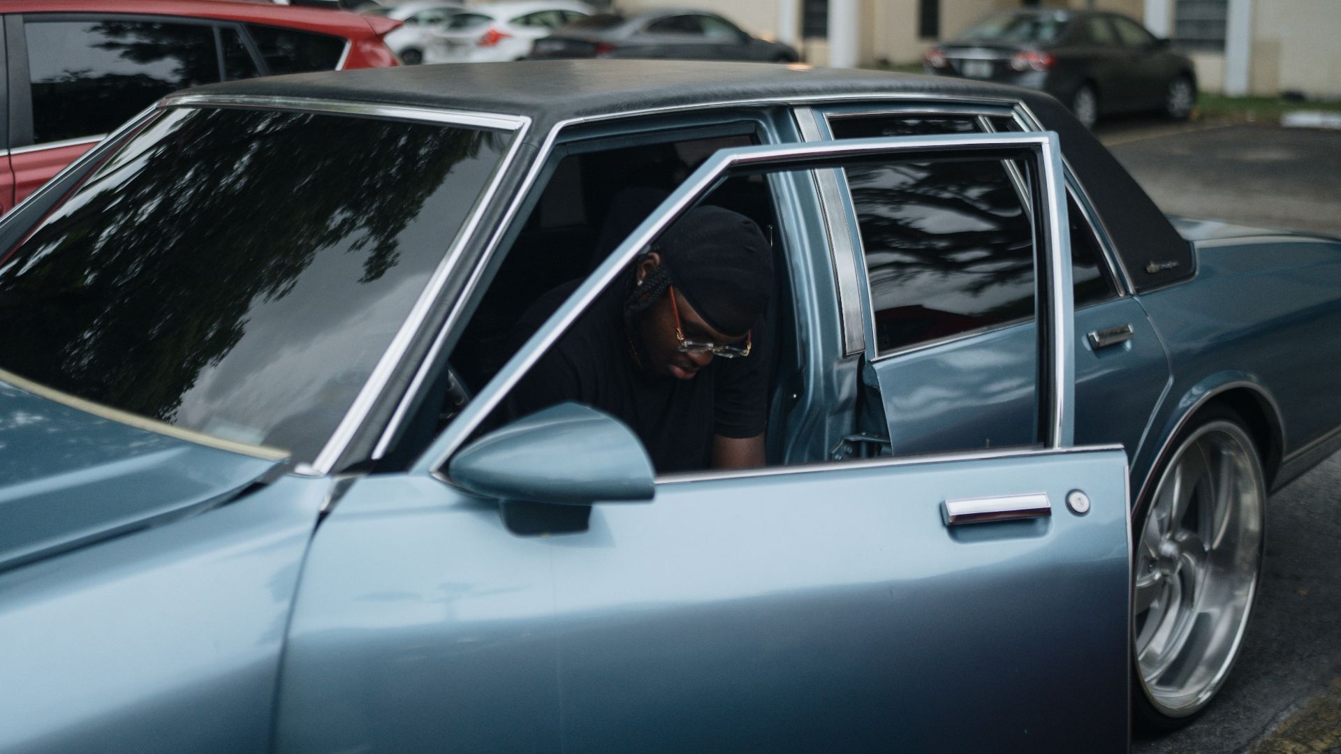 A man sitting inside a parked car with an open door in a parking lot.