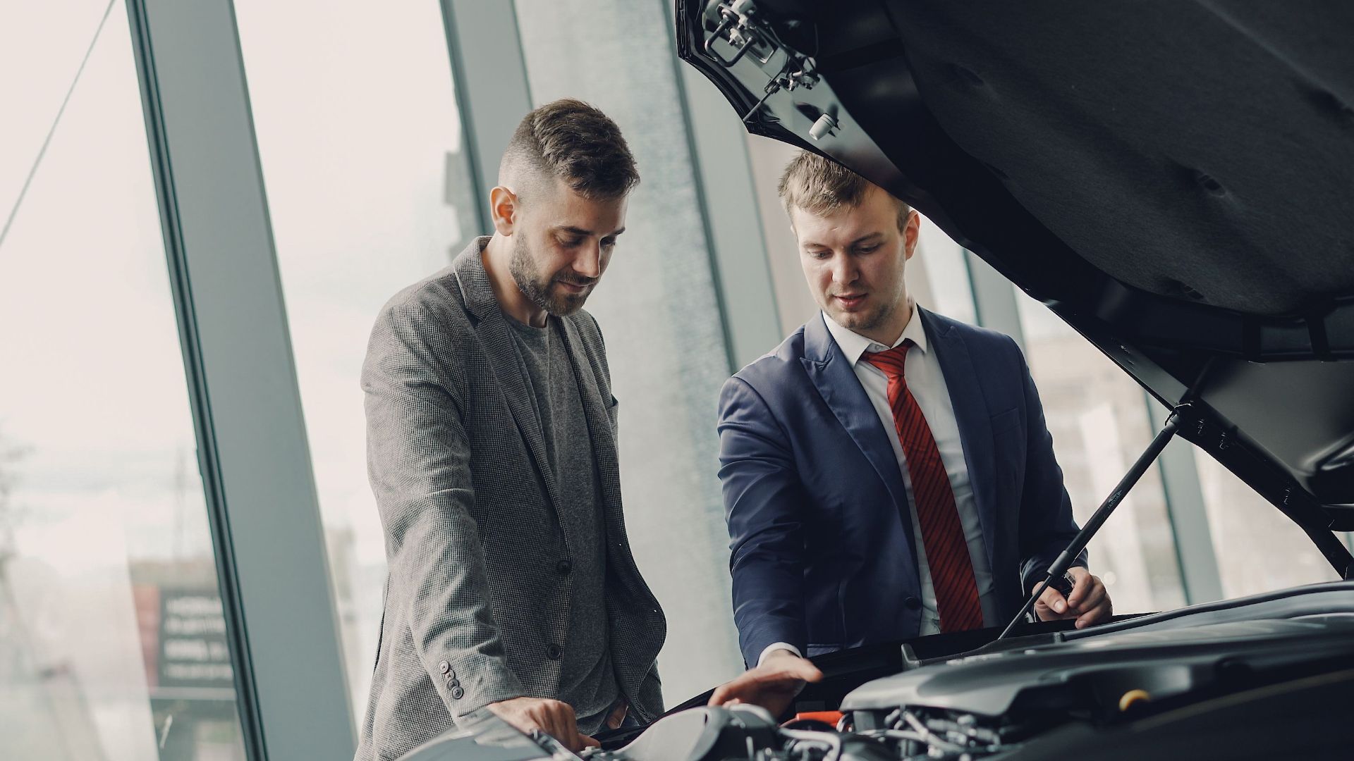 Two men inspecting a car's engine hood in a dealership showroom.