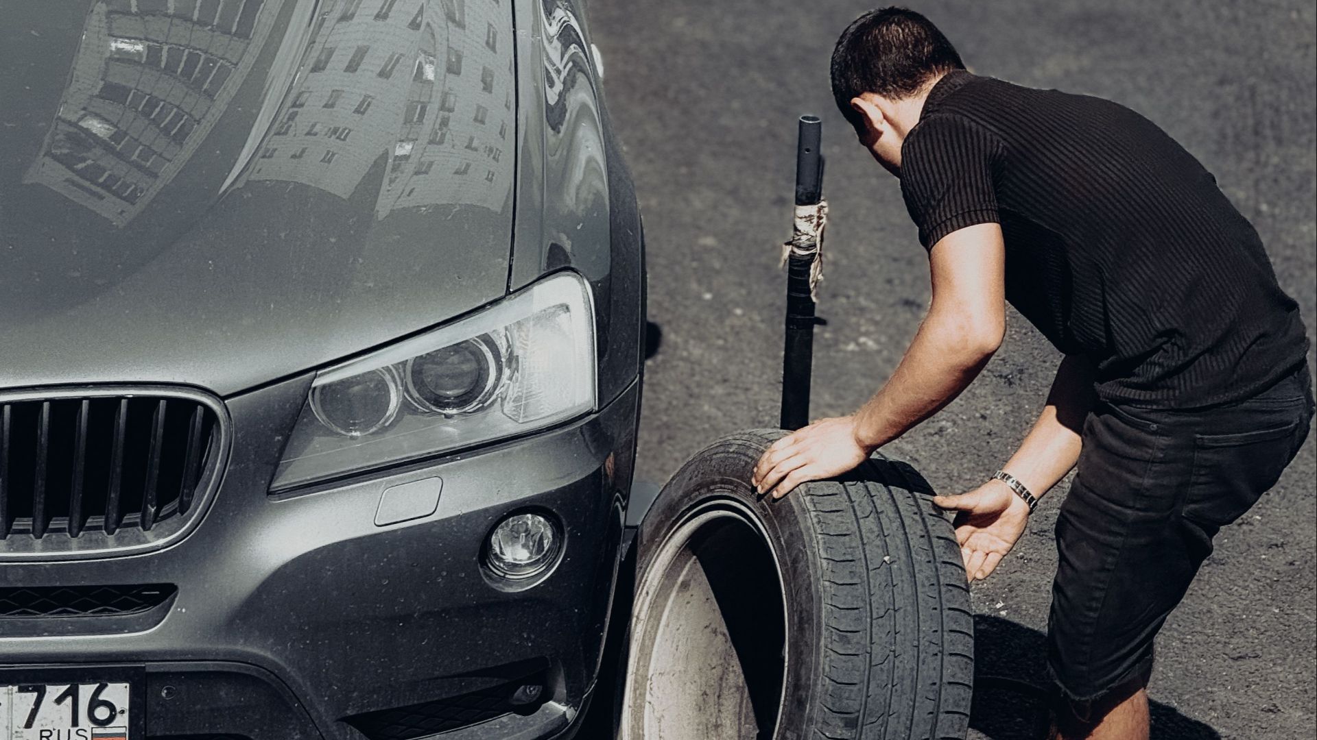 A young man changes a car tire on an outdoor road under the bright sunlight, highlighting the DIY task.