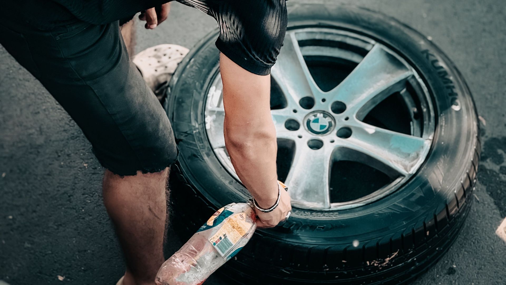 A person kneels on asphalt, cleaning a car tire with dedication, captured in natural daylight.