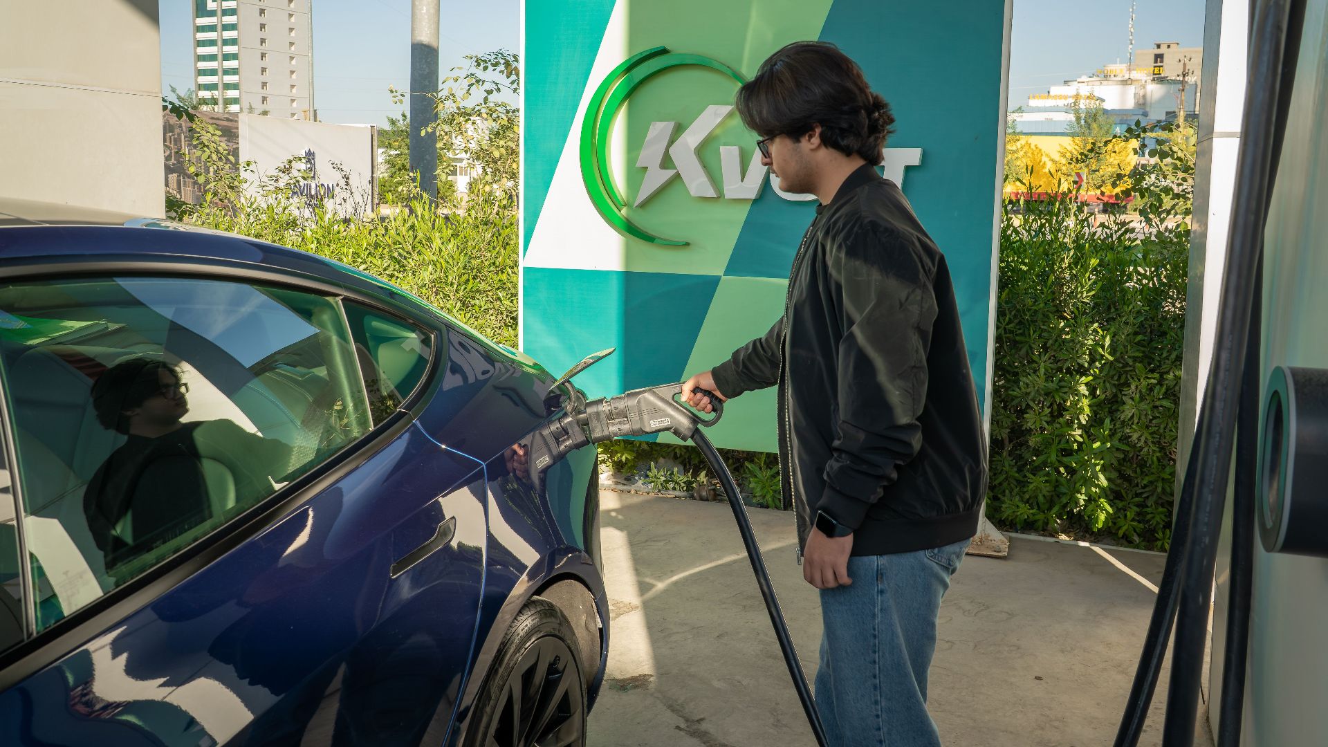 Young man charging a Tesla Model 3 at an outdoor electric vehicle station during the day.