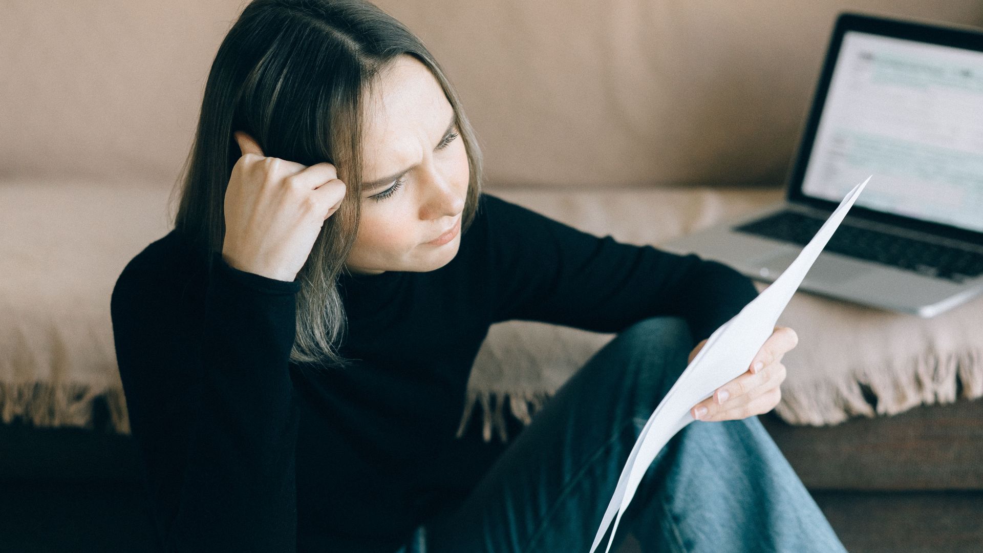 Focused woman analyzing papers with a laptop open, symbolizing thoughtful consideration.