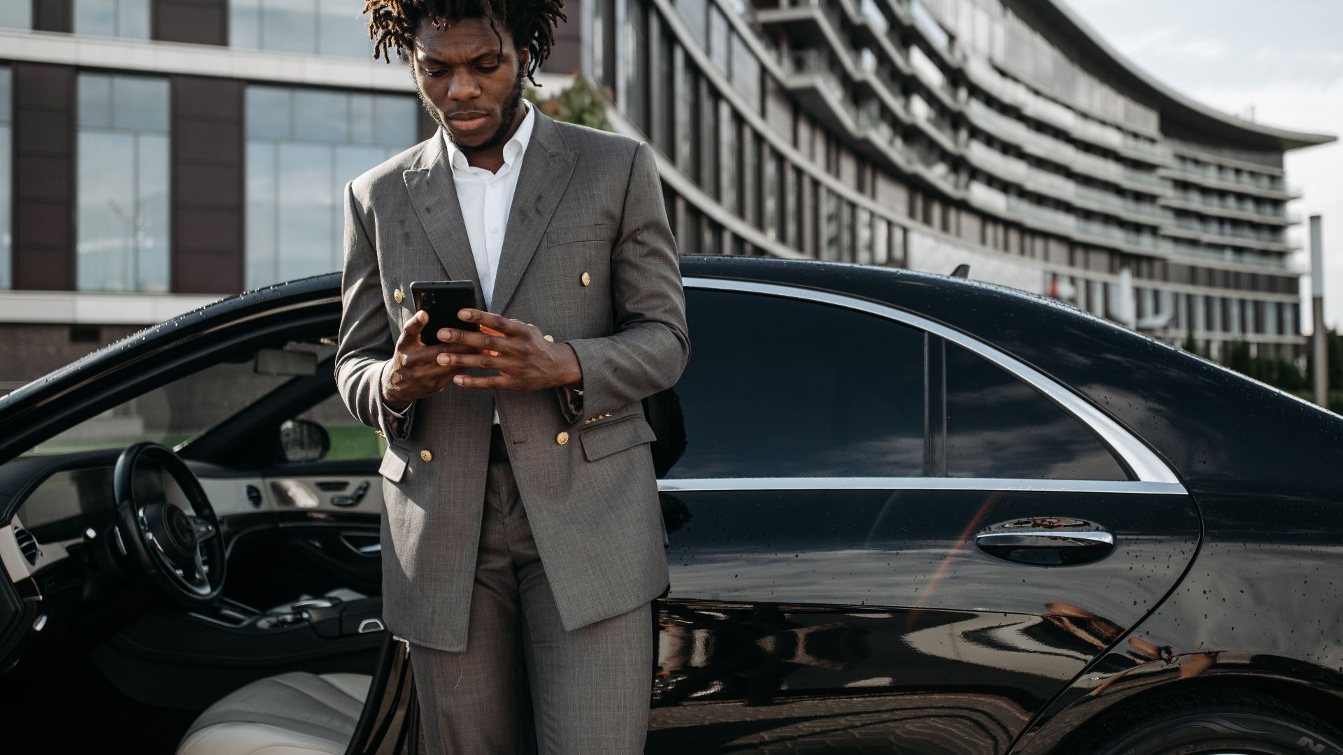 Professional man in a gray suit using his phone next to a luxury car.