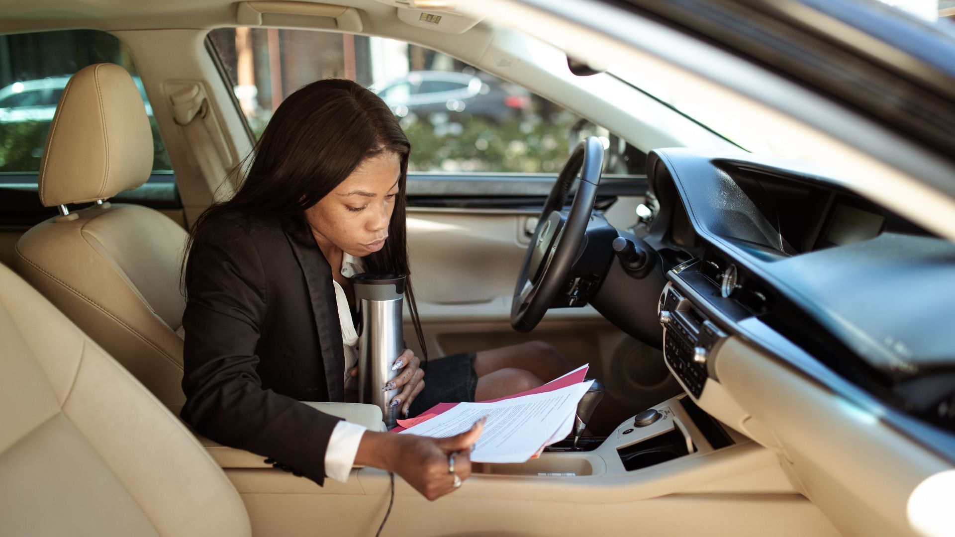 A focused businesswoman reads documents in her car, multitasking with a tumbler.