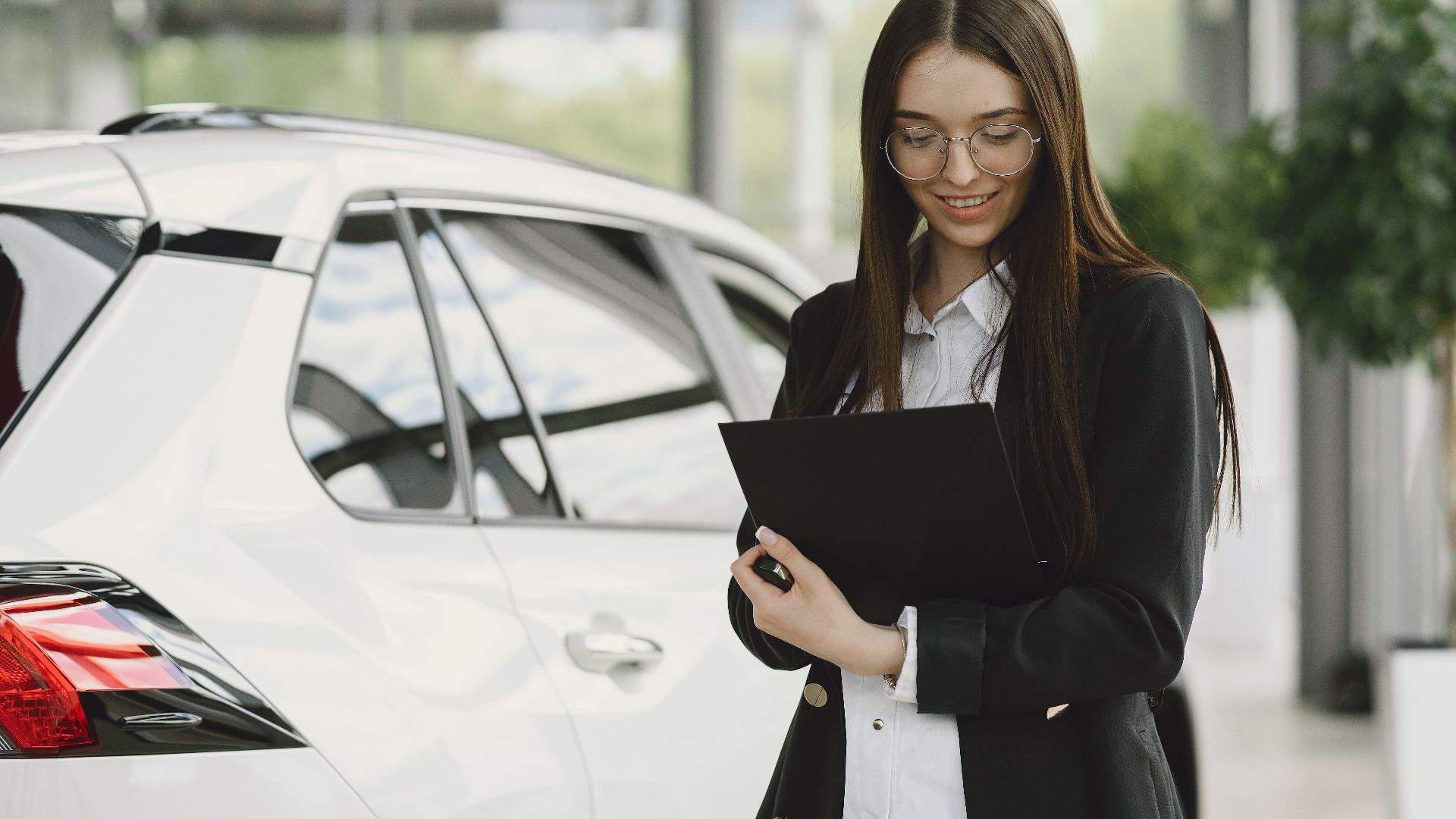 Smiling woman in a black jacket reviewing documents near a white car indoors.