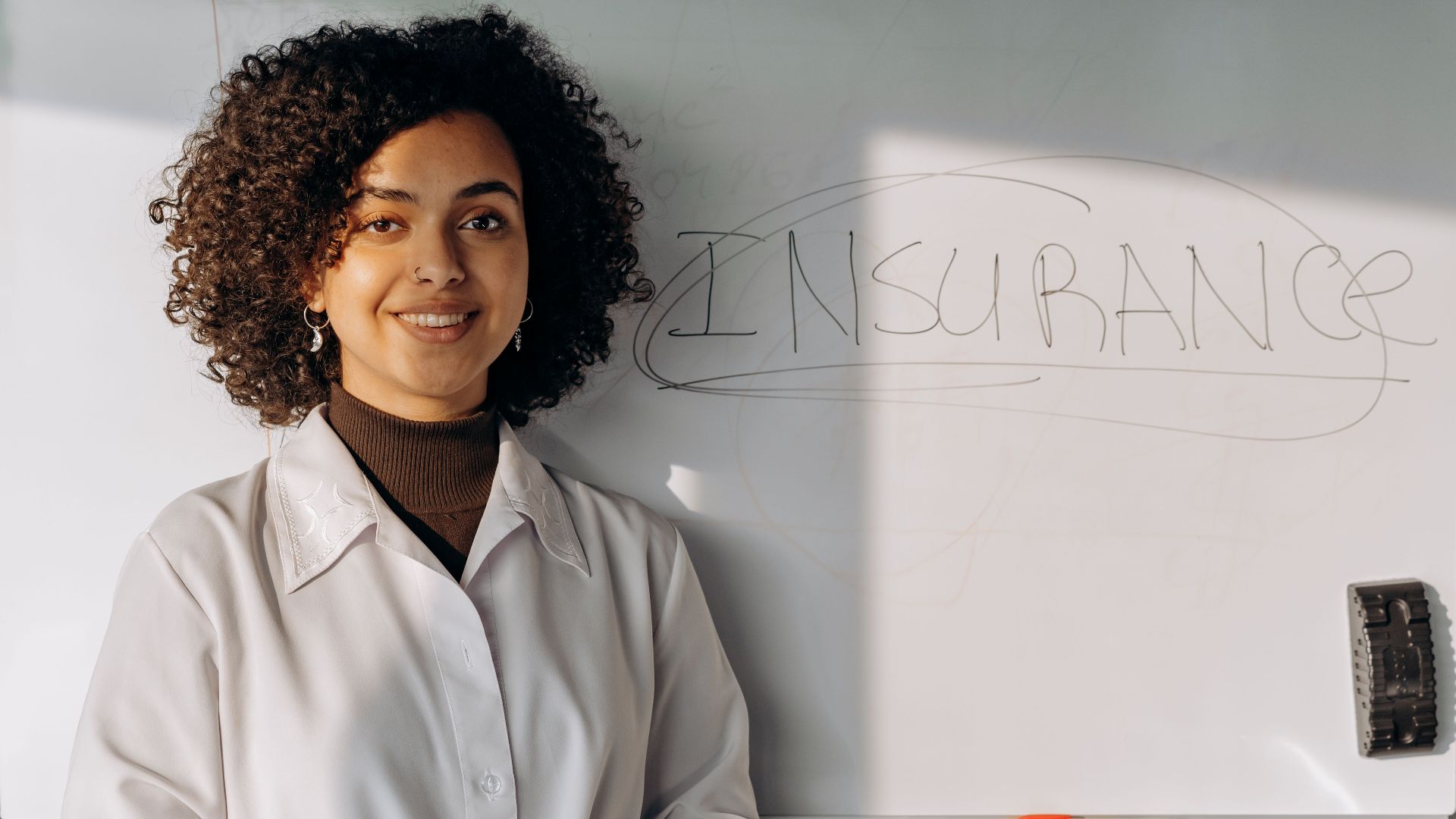 African American woman smiling in office setting with a whiteboard displaying 'Insurance'.