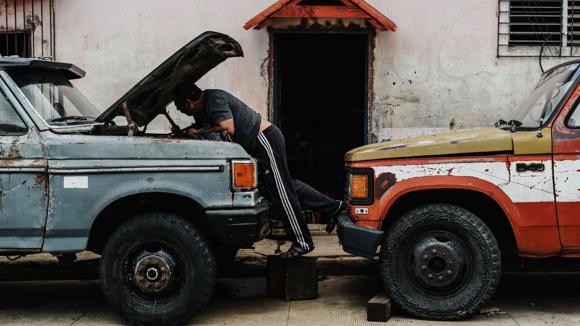 Man working on car engine outdoors, examining under the hood of classic trucks parked closely together.