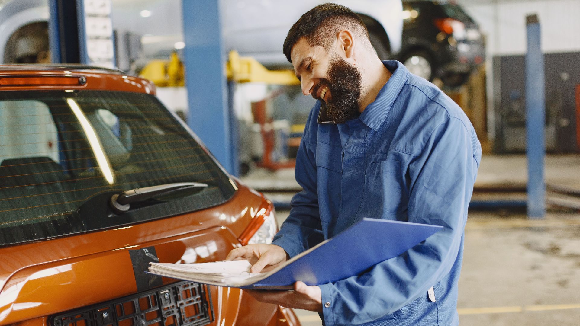 Smiling auto mechanic inspects a car in a service garage, wearing coveralls and holding documents.