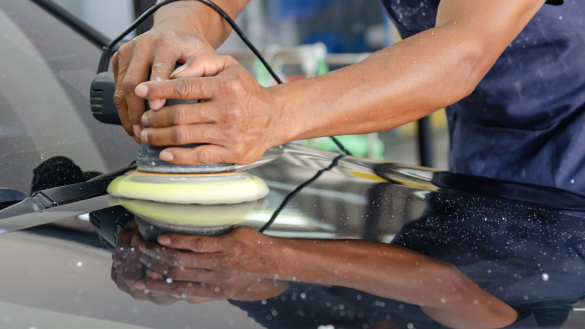 Detailed view of a worker's hands polishing a car's hood using a buffer in an auto workshop.