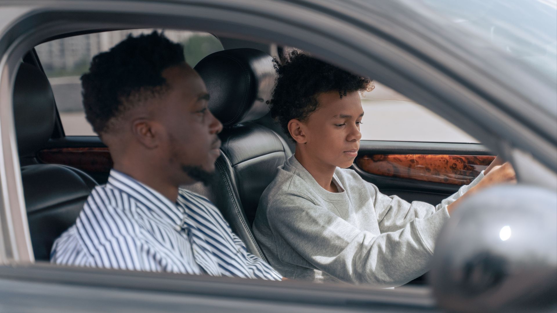 A father sits beside his son who is learning to drive, inside a car.